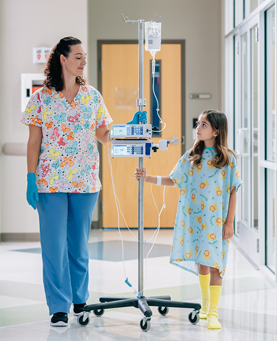 Nurse is walking down hallway, guiding pole holding infusion pumps and a Baxter premixed solution bag; pediatric patient receiving the infusion walking next to nurse.