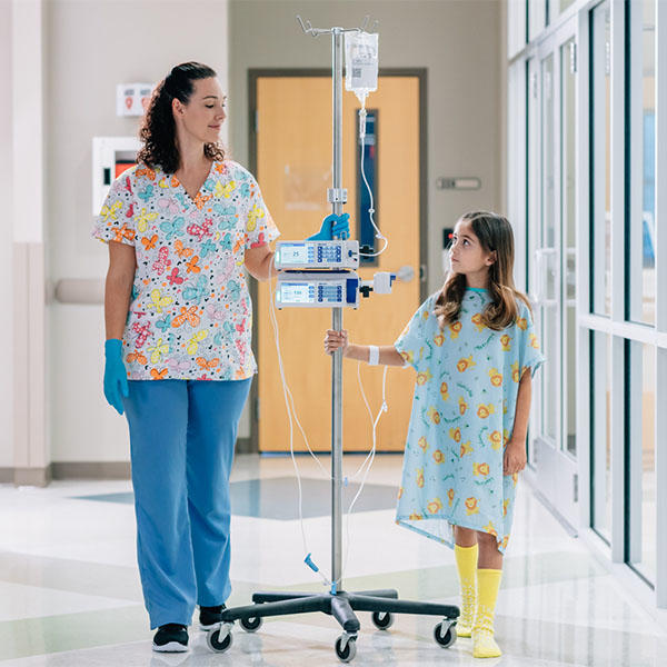Nurse is walking down hallway, guiding pole holding infusion pumps and a Baxter premixed solution bag; pediatric patient receiving the infusion walking next to nurse